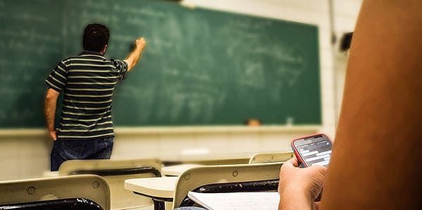 A man writing on a green chalkboard in front of student in classroom.