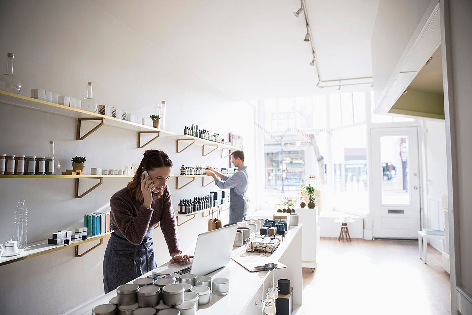 Female shop owner talking on cell phone and working at laptop in home fragrances shop